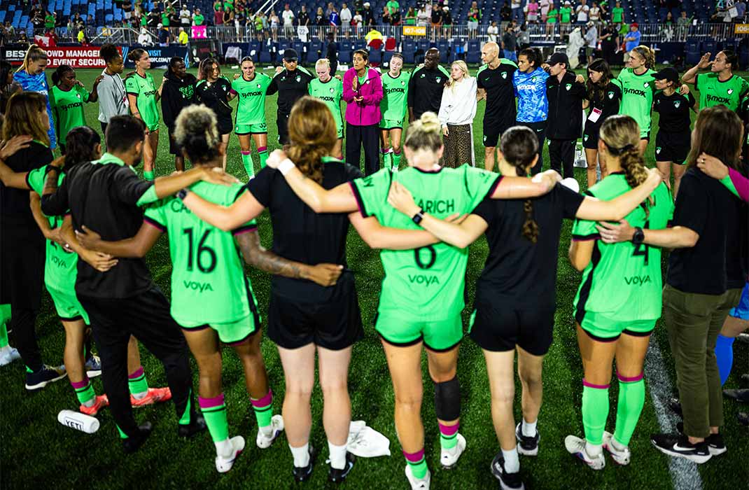 A women’s soccer team in bright green uniforms stands in a large huddle on the field after a match. These players from Boston Legacy FC link arms with coaches and staff, forming a circle that fills the foreground. Stadium lights illuminate the scene while spectators and additional team members are visible in the background.