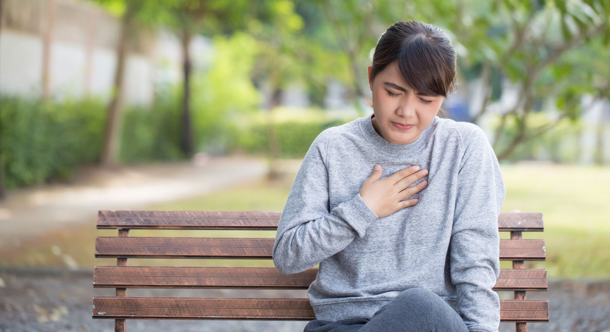 A woman on a park bench touches her chest, feeling heartburn pain from GERD.