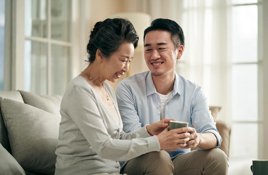 A son hands a mug to their mother as they sit together smiling on the couch
