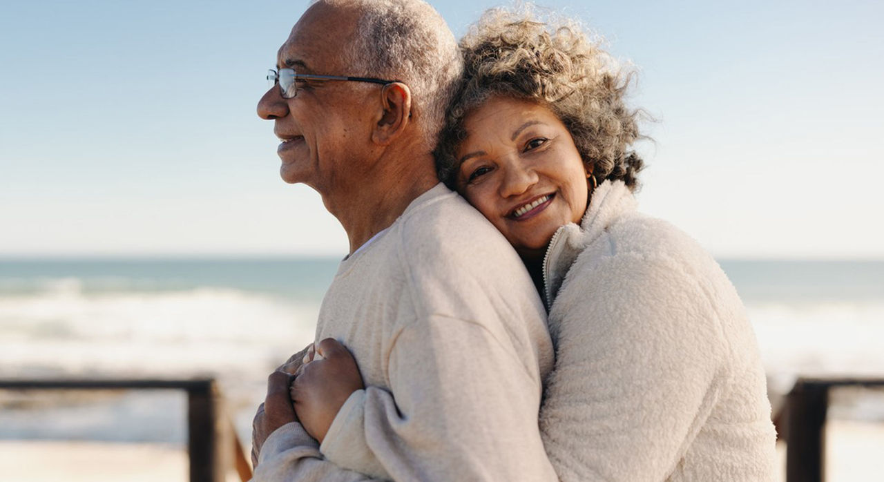 A smiling woman hugs her partner from behind at the beach
