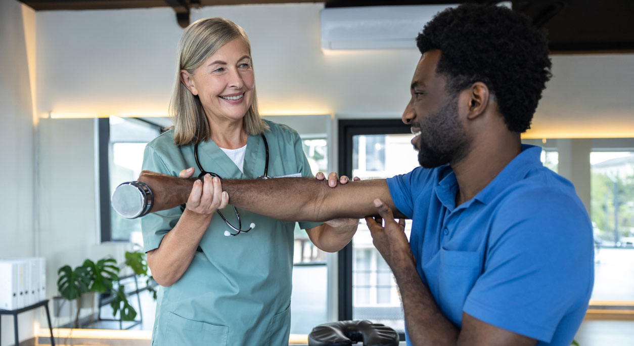 A physical therapist guides a young man in lifting a dumbbell with one hand.