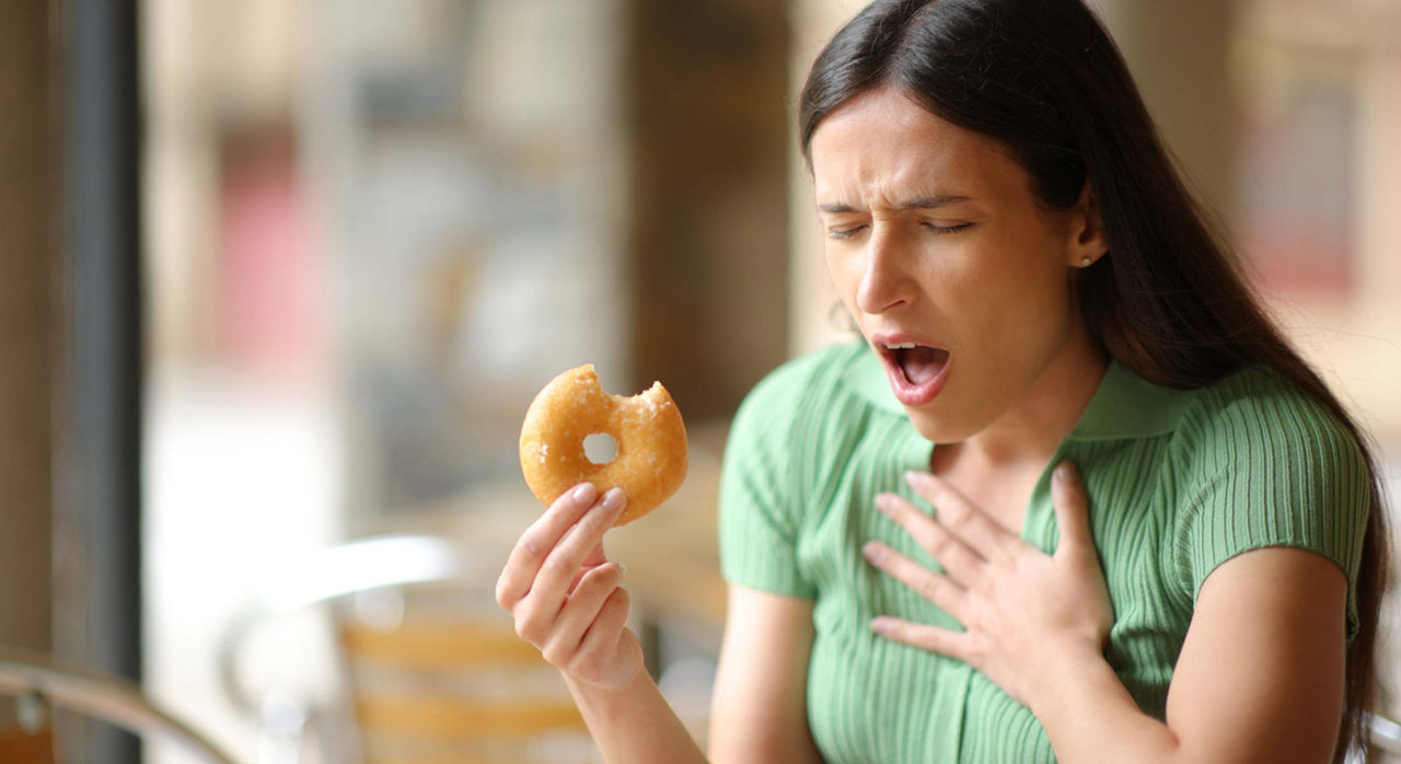 A woman choking with her left hand on her chest after eating a doughnut that she's holding in her right hand