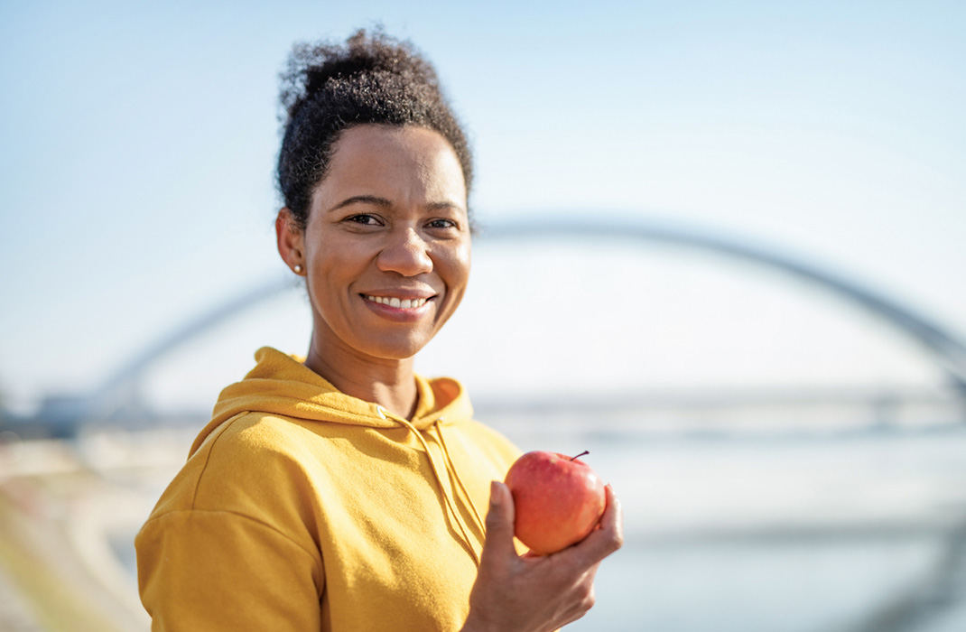 A woman wearing a mustard hoodie with her hair tied back smiles and holds a red apple in right hand