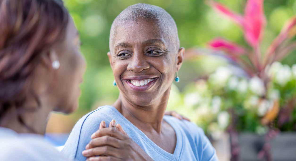 A smiling older woman holds her friend's hand supportively.