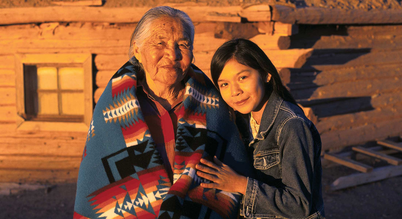 A Native American grandmother and granddaughter in front of a traditional house.