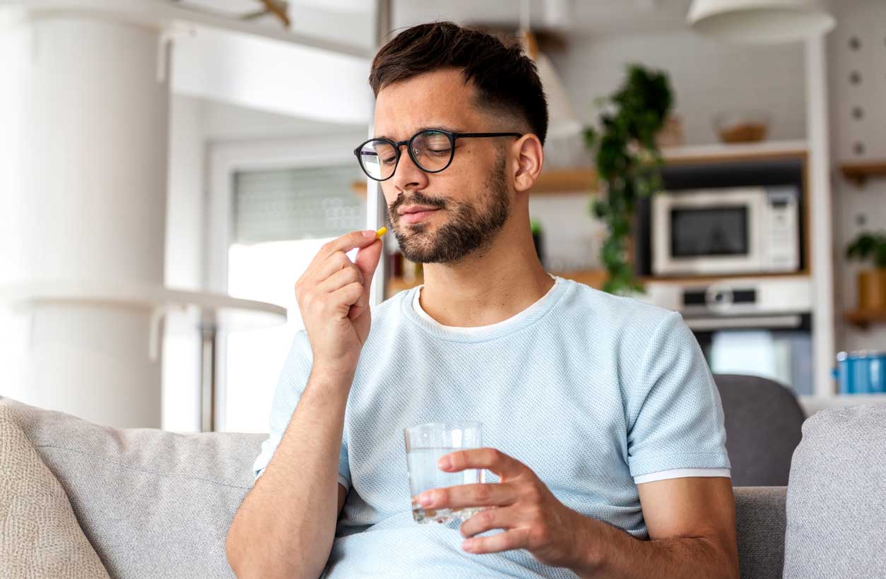 A bearded young man wearing glasses and a short-sleeved shirt about to take a prescription pill, holding the pill in one hand and a glass of water in the other.