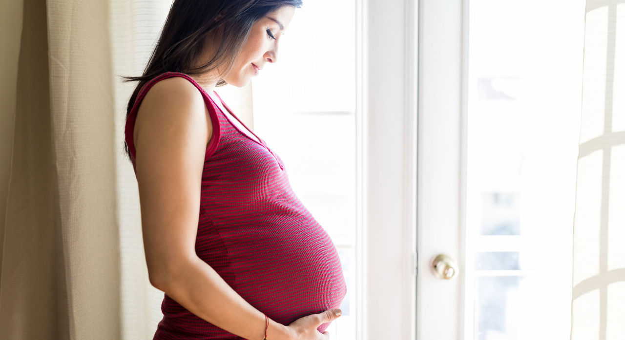 A heavily pregnant woman stands by a window, cradling her belly.