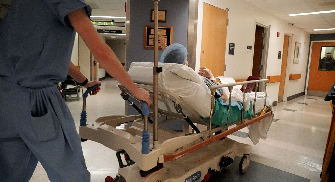 A medical professional in blue scrubs pushes a patient on a hospital stretcher through a brightly lit corridor. The patient is covered with a blanket and wears a surgical cap.