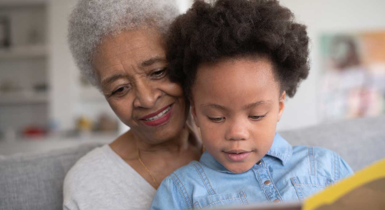 A grandmother reads with her toddler grandson, who sits in her lap.