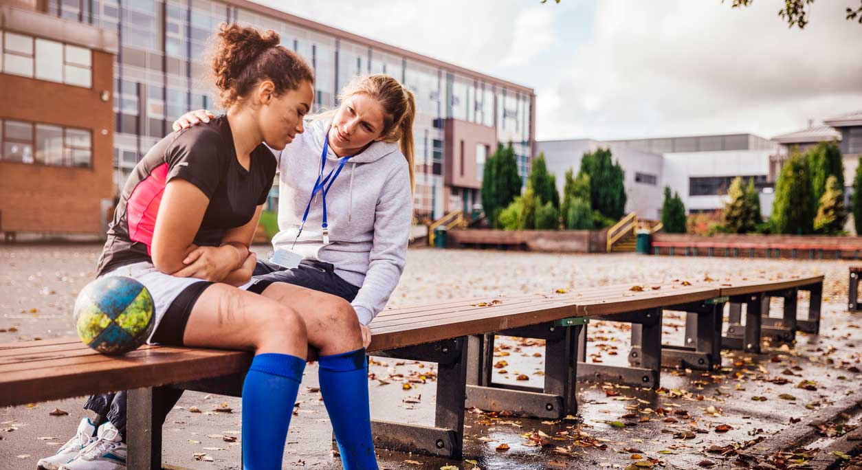 A coach comforts a female athlete on a bench in a high school courtyard.