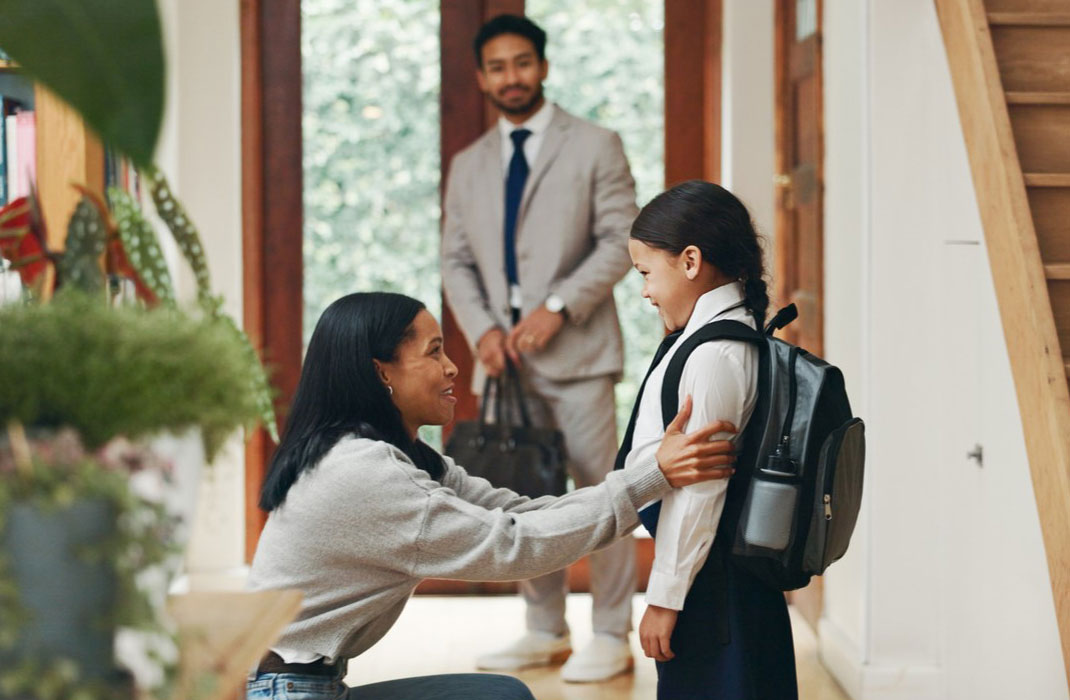A mother and father smile at their young daughter by their front door, ready to leave for her first day of school.