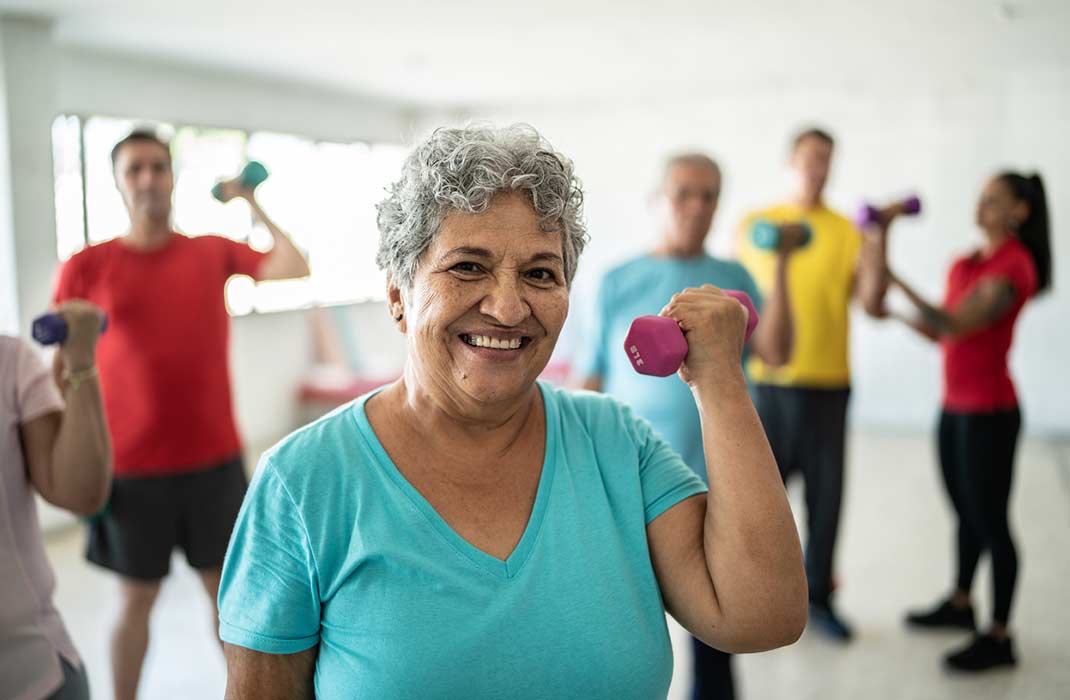 An older woman with white hair in a light teal t-shirt smiles while lifting up a pink hand weight with her left hand. There are 4 to 5 people in the background also lifting hand weights.