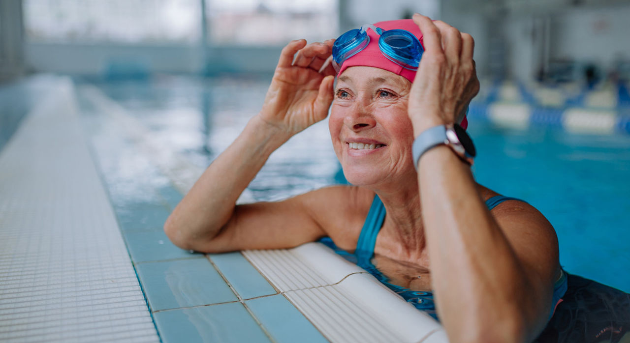 A smiling older woman wearing a teal swimsuit in a pool as she rests her elbows on the edge of the pool. Both of her hands, with a watch on her left hand, are on her red swimming cap that she is wearing while blue goggles sit on her forehead.