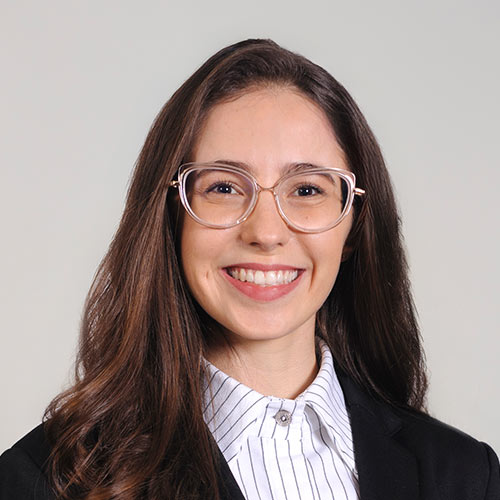 A woman with eyeglasses and long brown hair is smiling and dressed in a black blazer and a white pinstripe shirt.