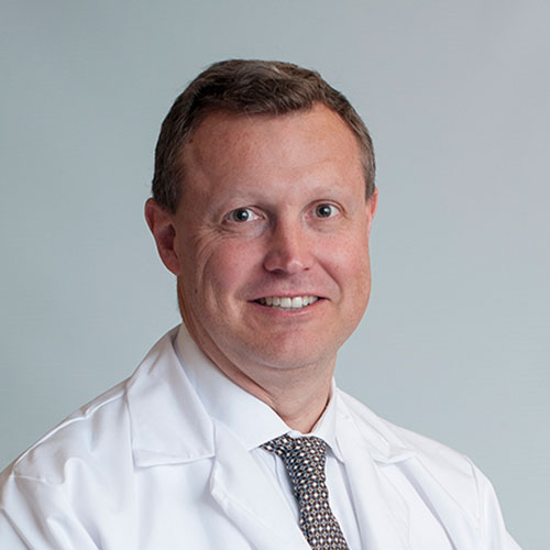 A man wearing a white shirt and white coat with a patterned tie is posed for a headshot against a neutral background