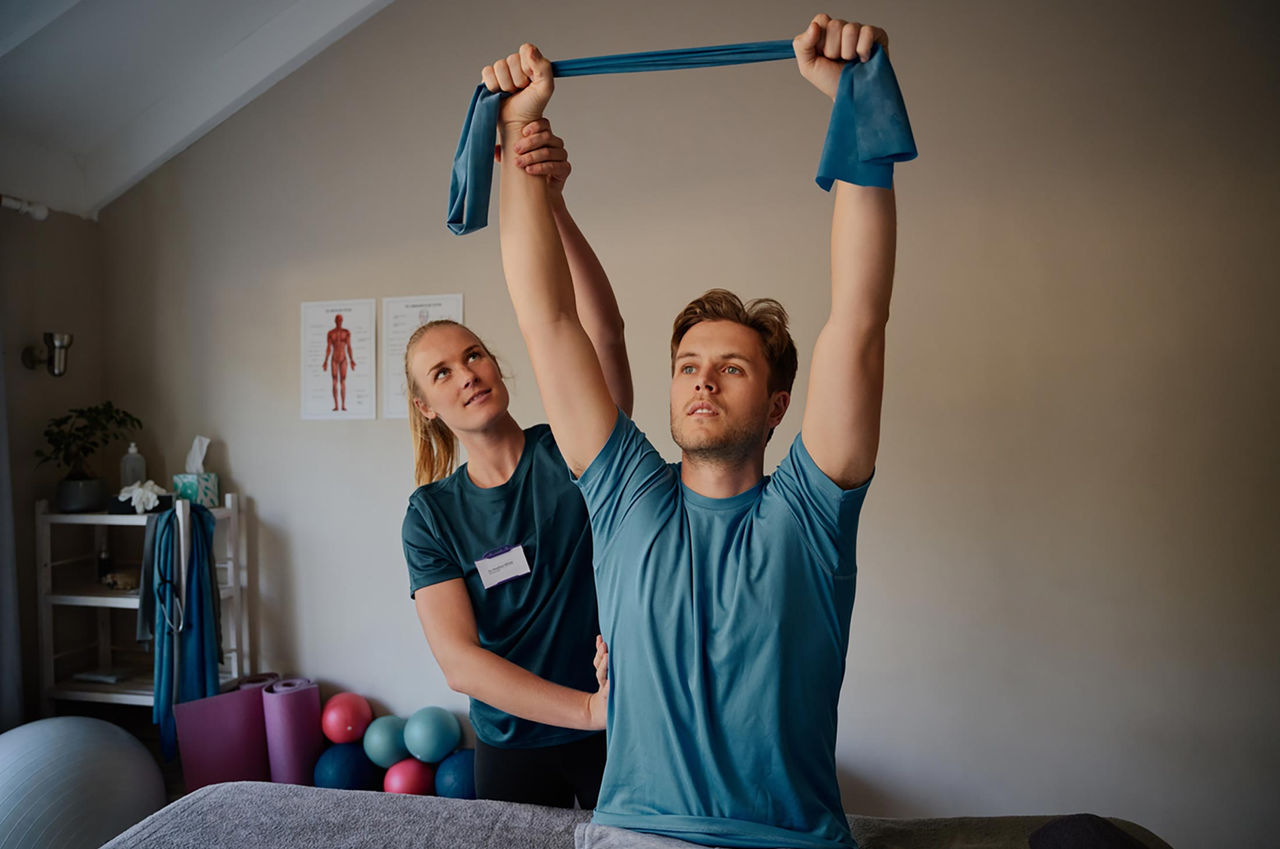 A young man uses a resistance band above his head as a physical therapist corrects his form