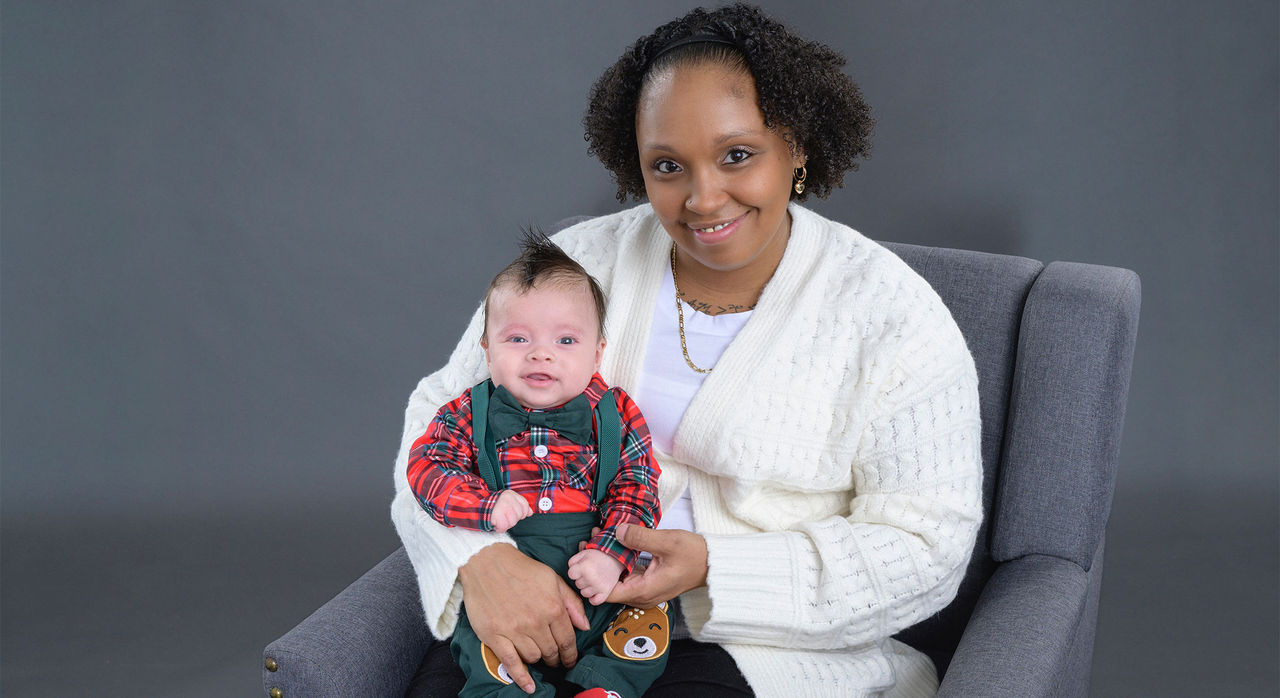 A mother holds her infant son in a studio photoshoot set against a gray background.