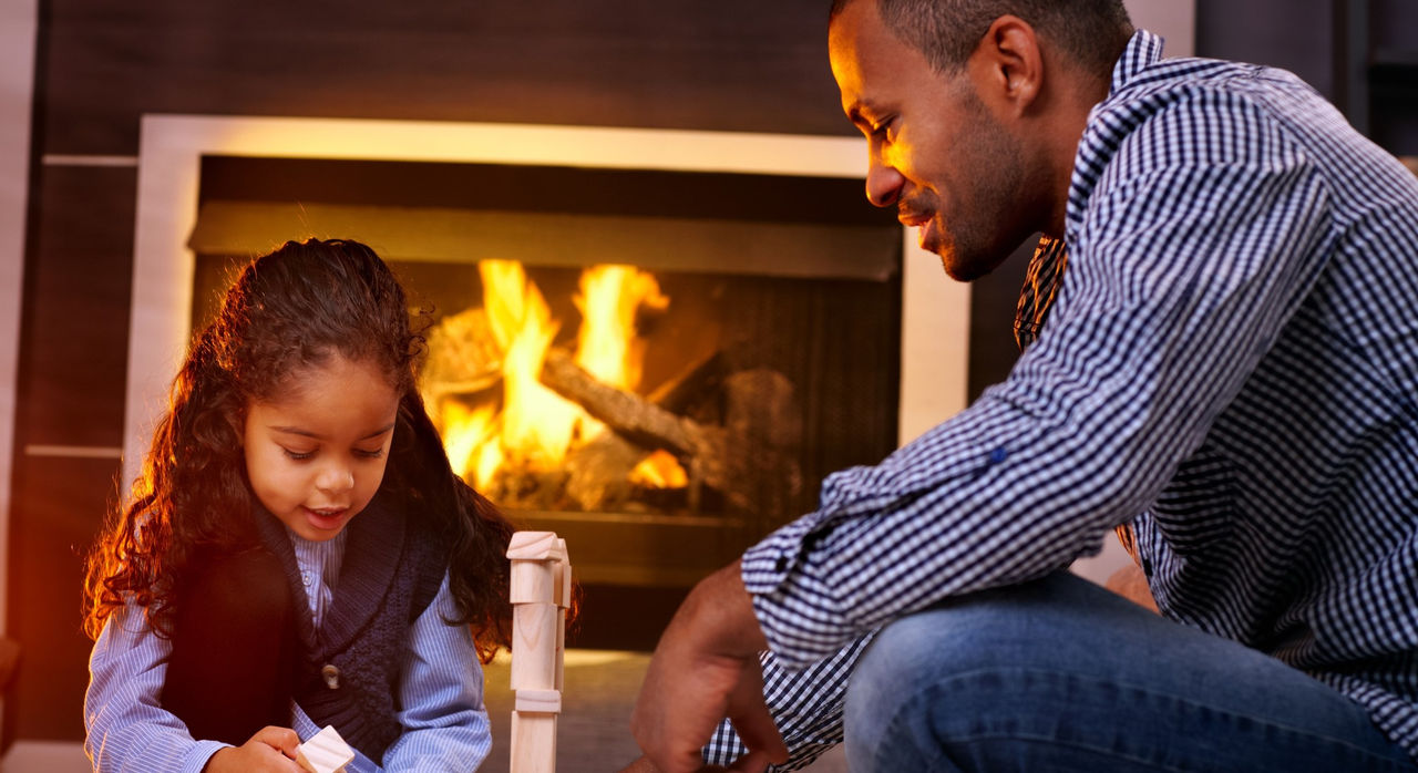 A child and her father play in front of a fireplace.