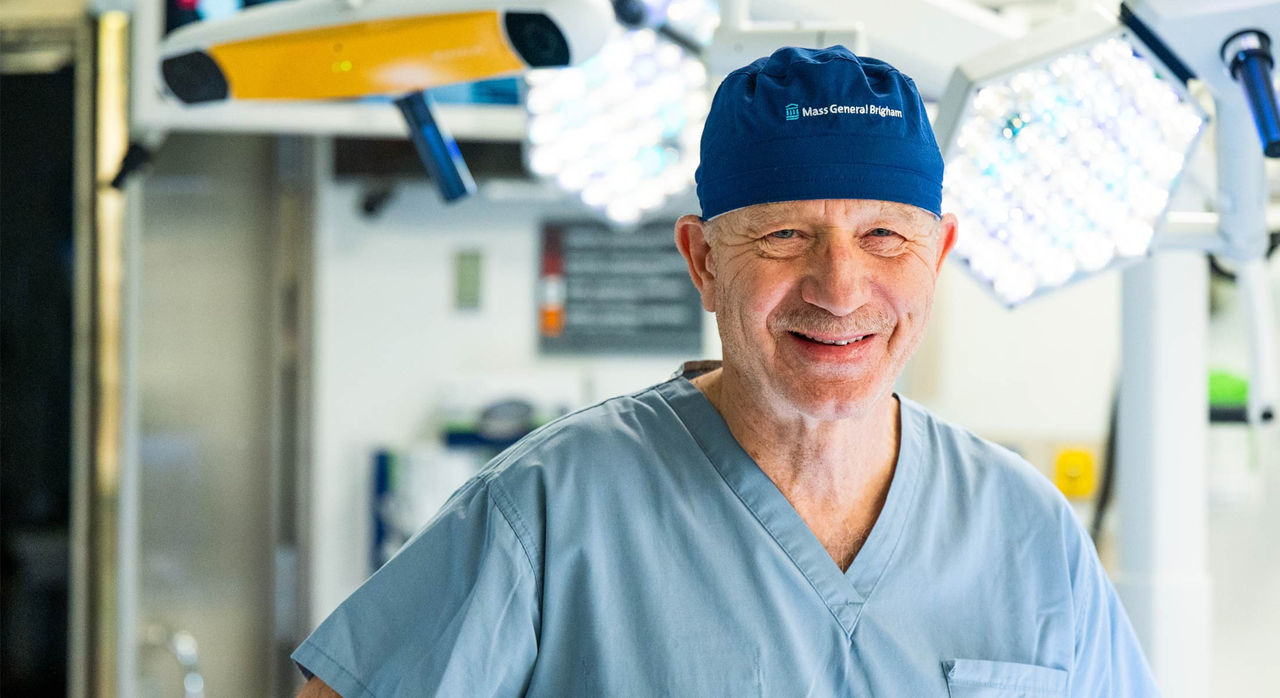 A surgeon in blue scrubs and a dark scrub cap with the Mass General Brigham logo stands and smiles in an operating room
