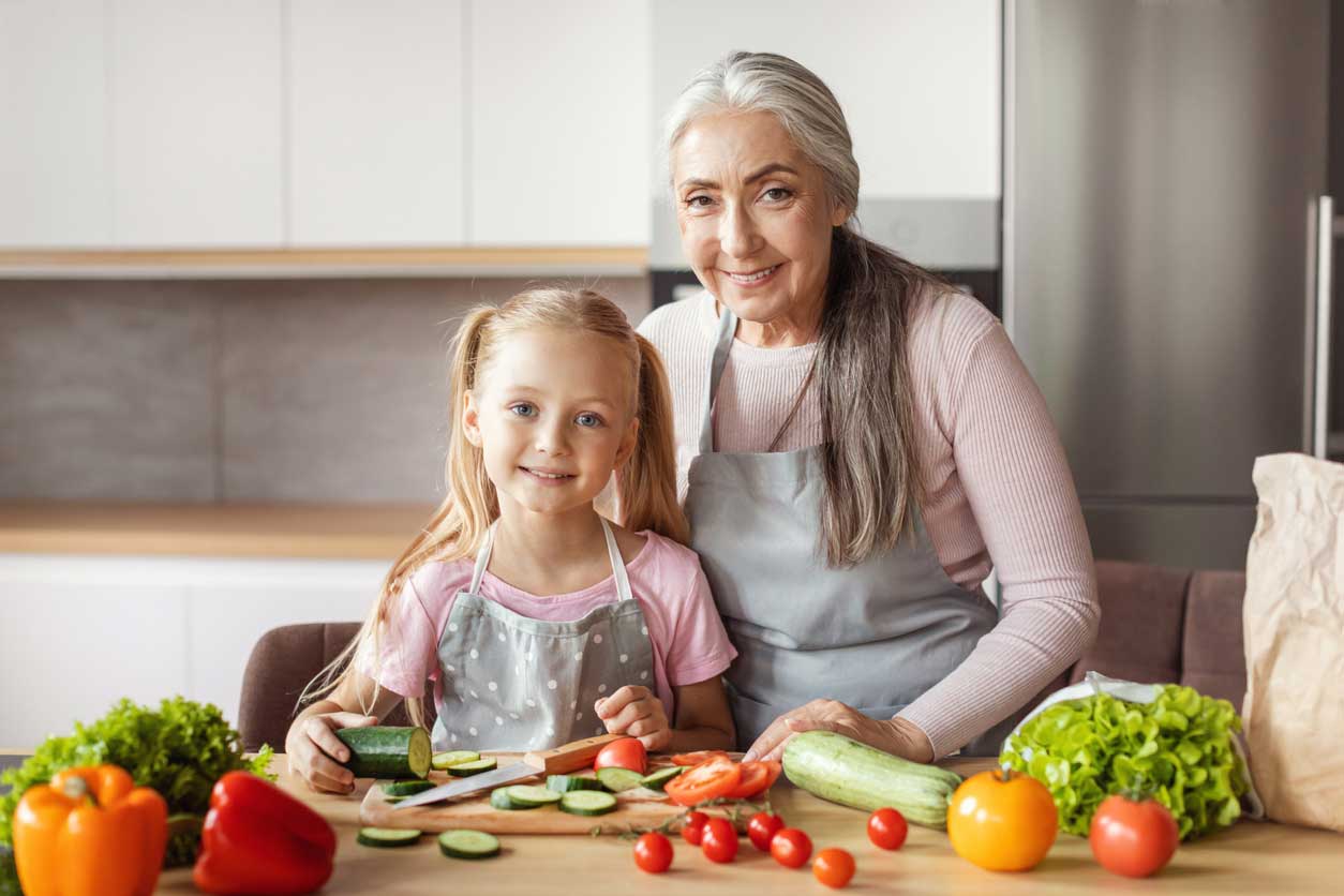 Grandma and granddaughter in the kitchen, chopping tomatoes, bell peppers, cucumbers, and greens.