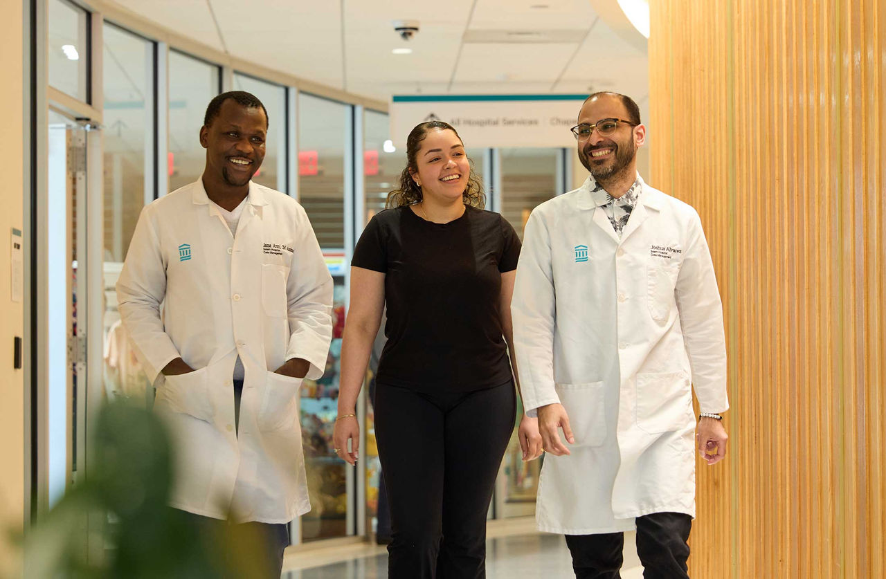Two smiling doctors in white coats walk with a smiling female patient down a hospital hallway.