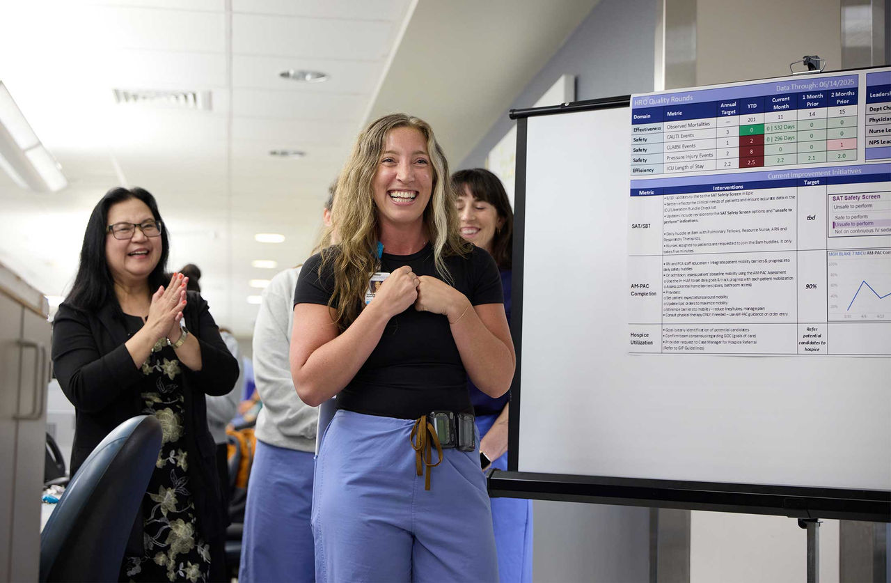 A nurse standing beside a presentation board smiles giddily as the people at her stand-up meeting applaud her presentation.