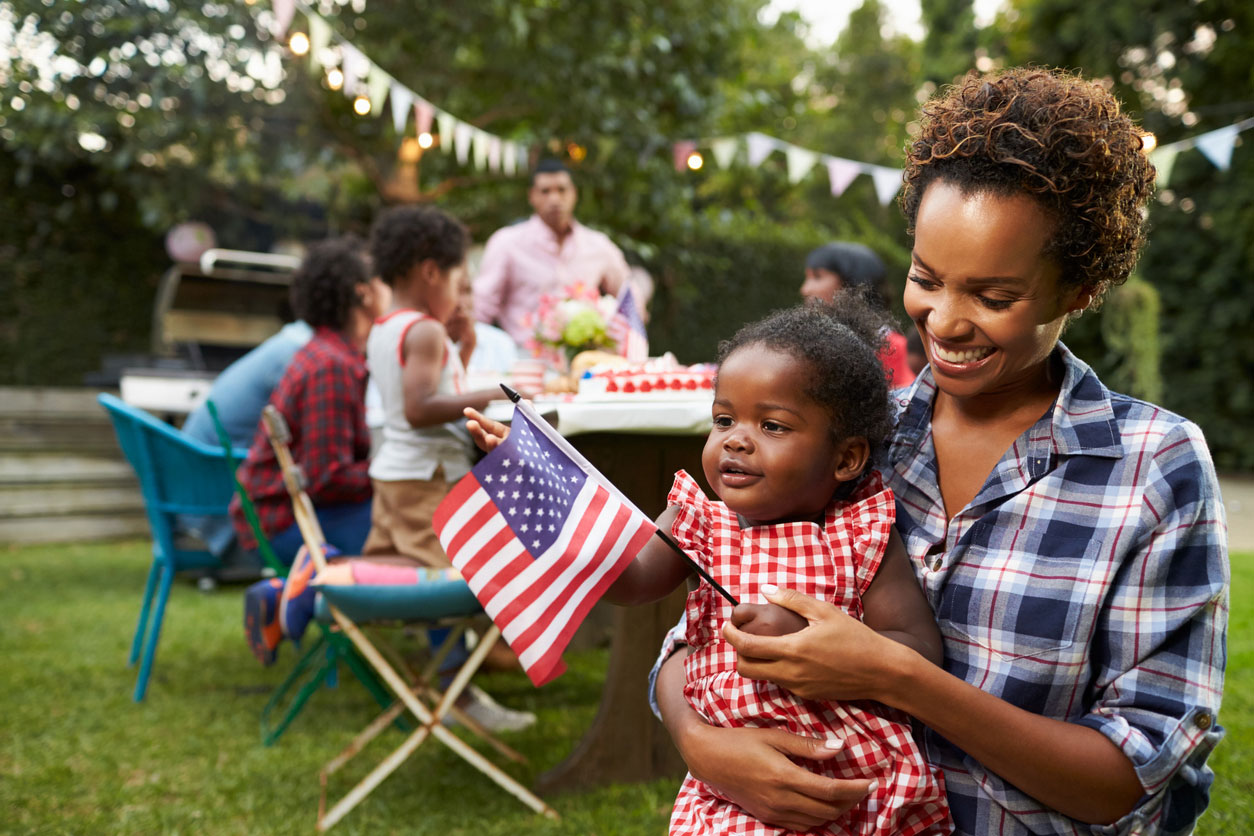 A smiling mom holds her young daughter and an American flag at a 4th of July backyard cookout.