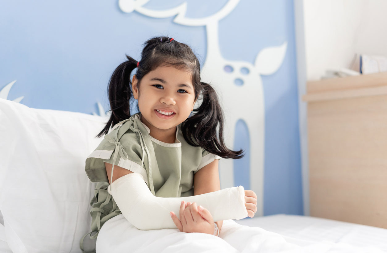 A smiling little girl with a cast on her arm sits in a hospital bed.
