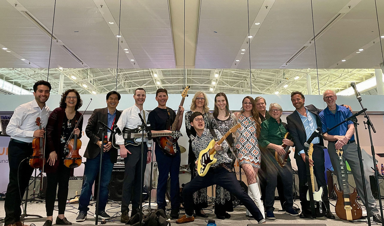 A group of more than 12 musicians smiling and posing together with their instruments like violins, guitars, basses, and more.