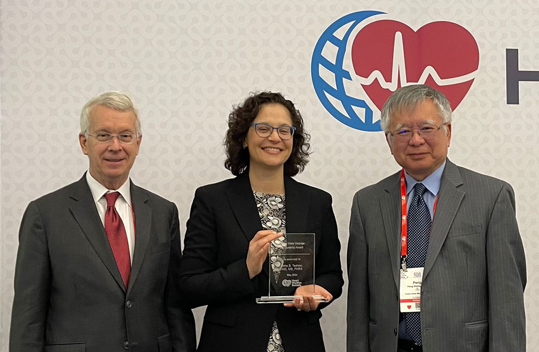 A man with white hair, white shirt, in a grey suit with a red tie stands next to a woman in glasses, a floral dress, and black blazer while she holds an award; next to her is a man with white hair, red lanyard, blue shirt and tie with a grey blazer.