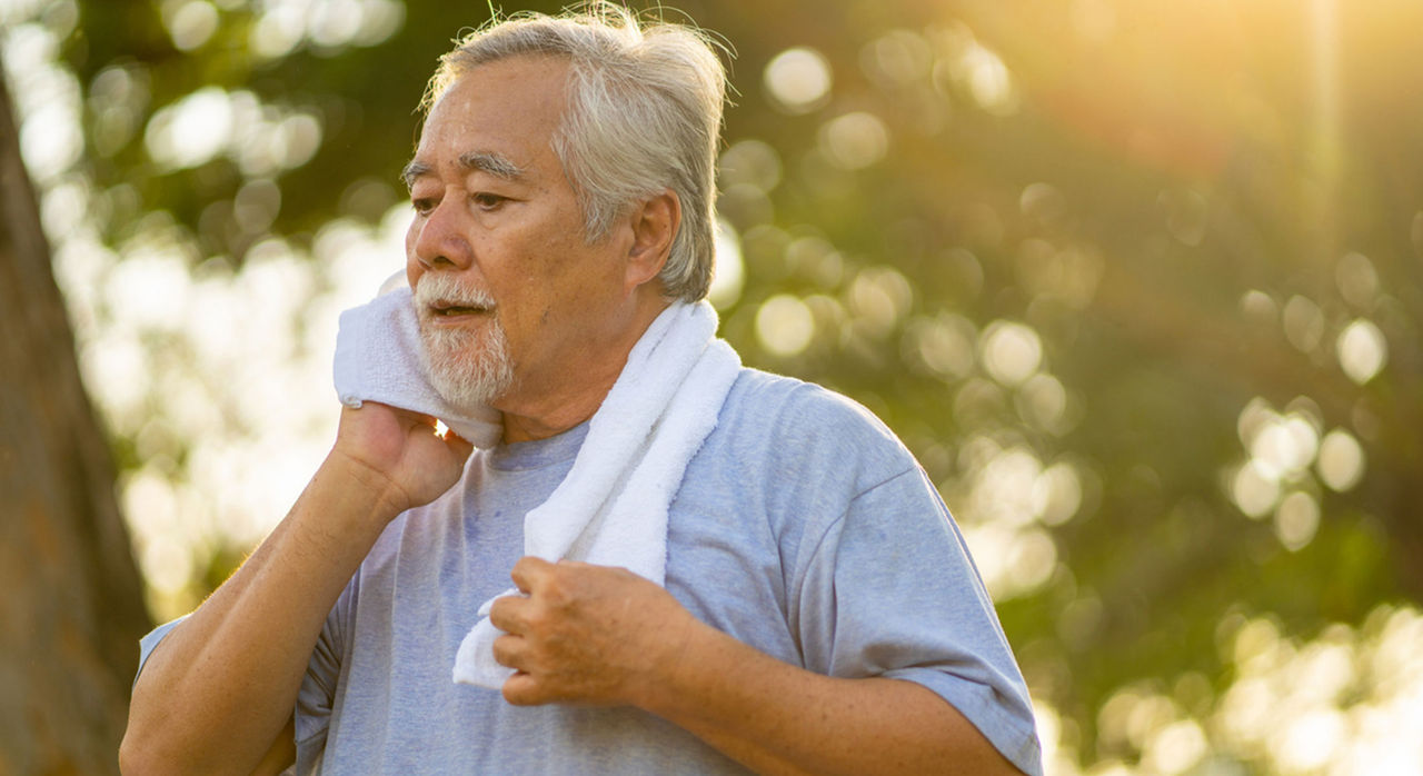 An elderly man uses a towel to wipe sweat from his face in a sunny park.