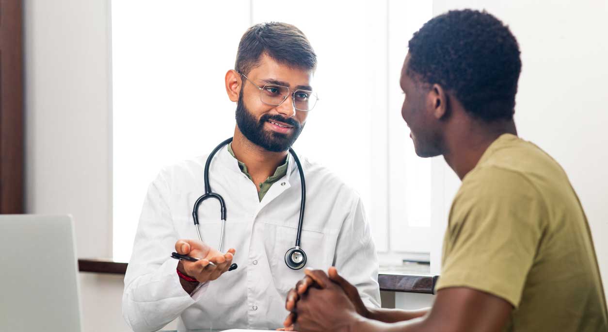 A doctor, with his right arm on his chest, chats with a patient in a medical setting; both are wearing masks