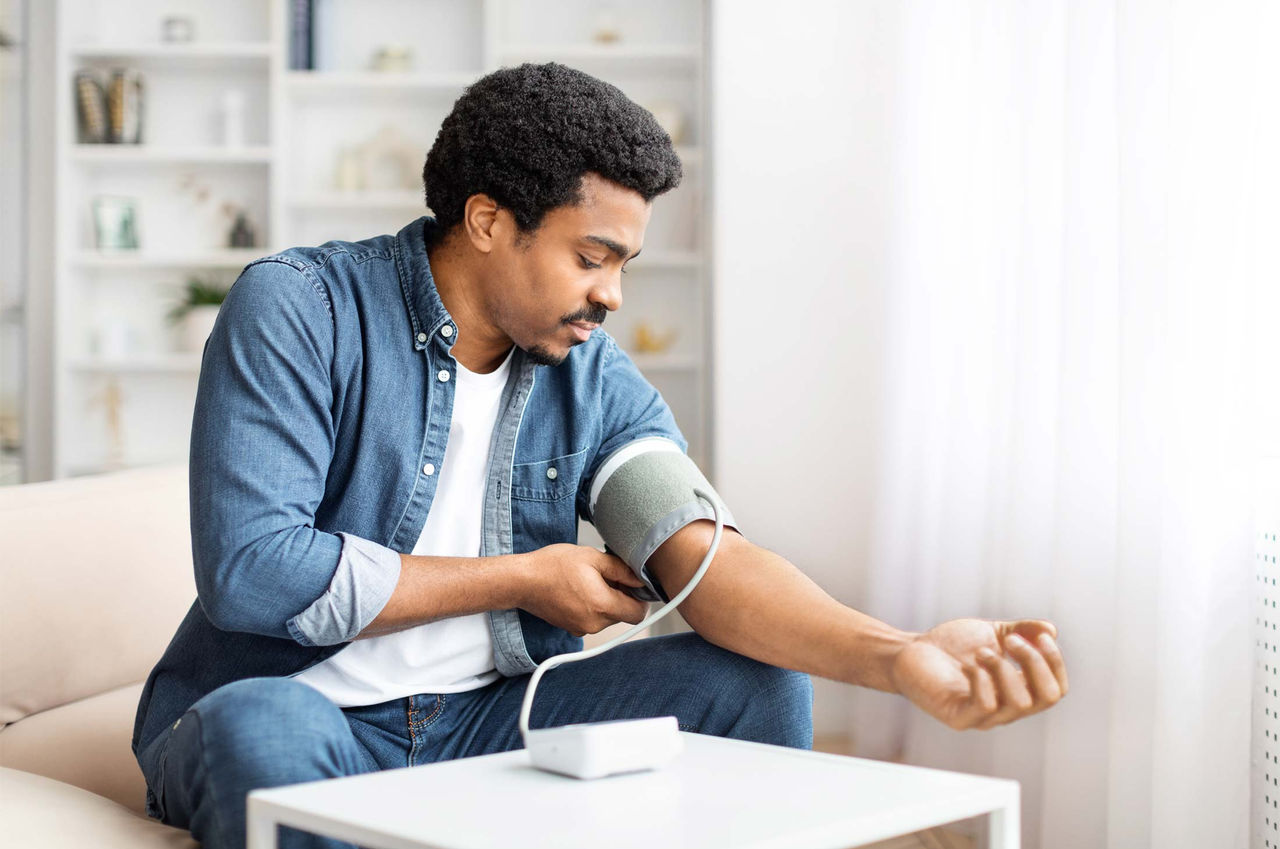 A Black man wearing an open blue denim shirt with a white undershirt and blue denim jeans monitors his blood pressure with a cuff while sitting on a couch