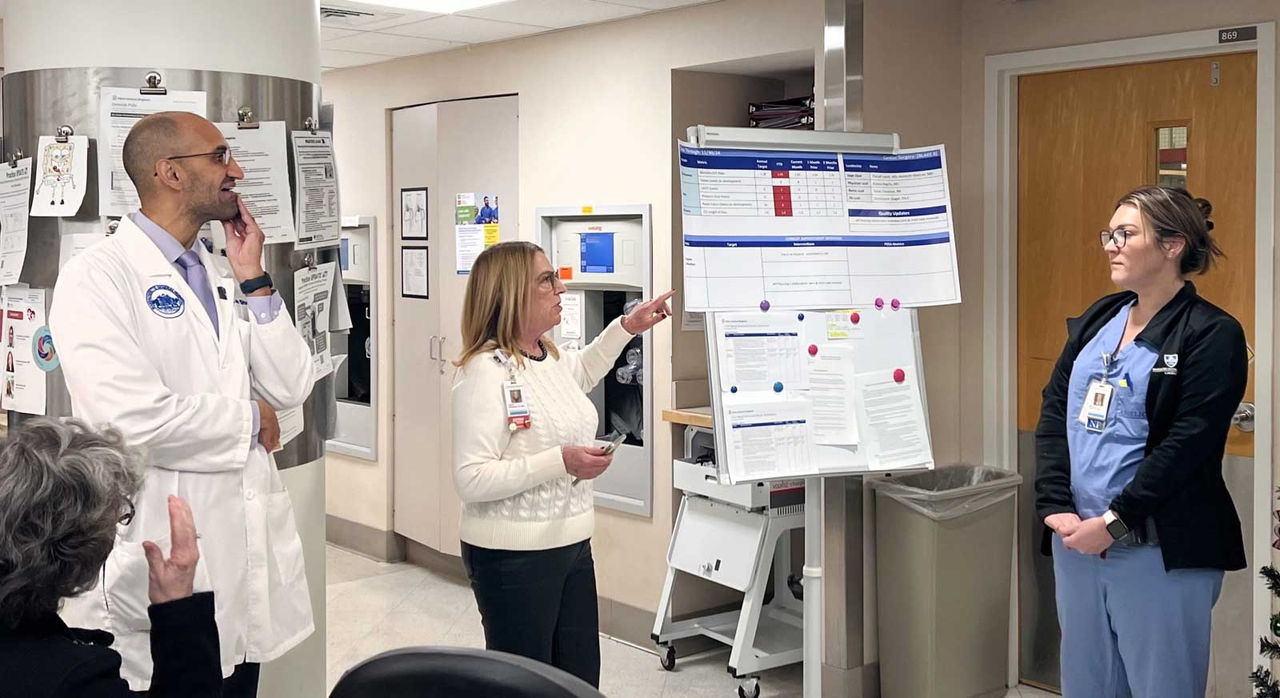 In a hospital hallway, a nursing director discusses papers pinned to a board with her team.