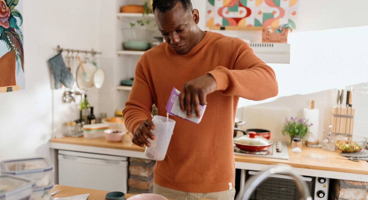 A man in an orange long-sleeve t-shirt preparing a protein shake in a kitchen