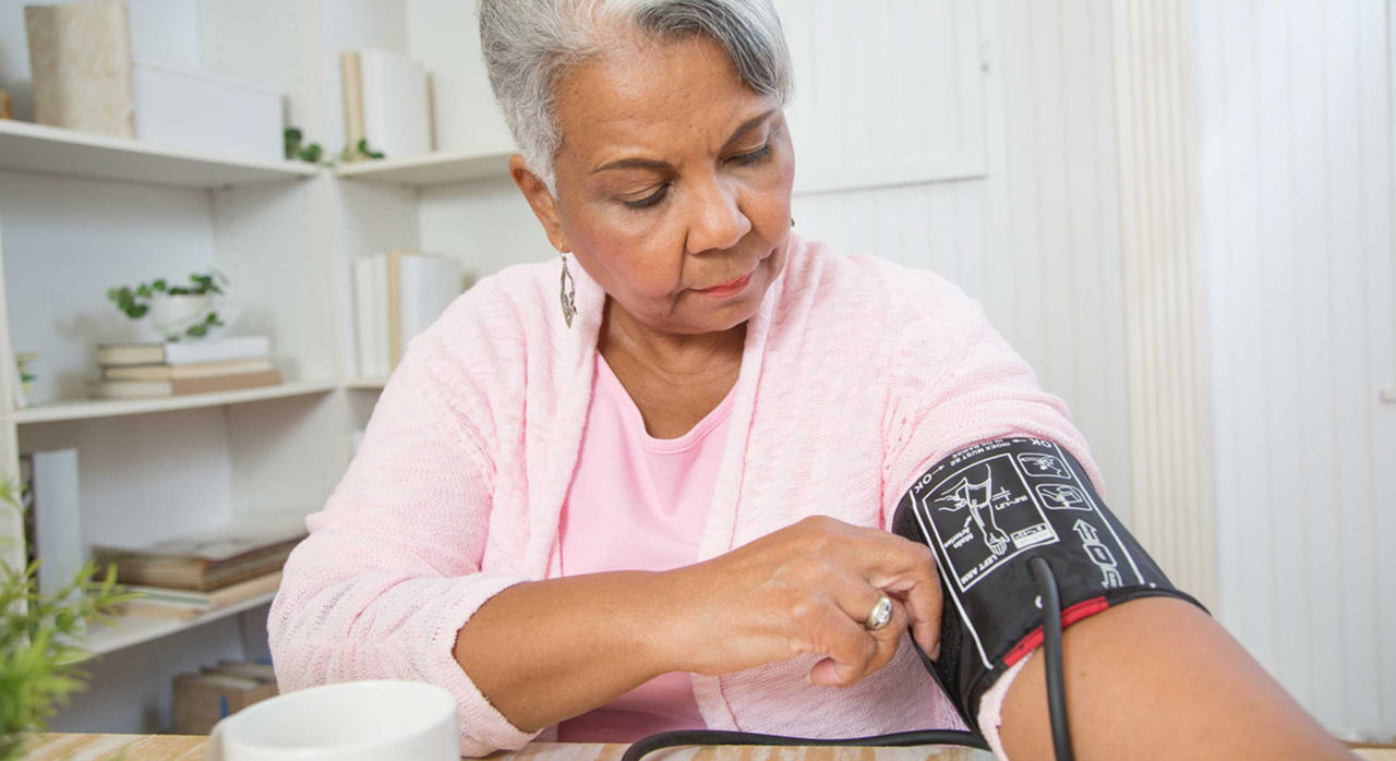 Woman with grey hair dressed in a pink top and cardigan uses a blood pressure cuff on her left arm to check levels