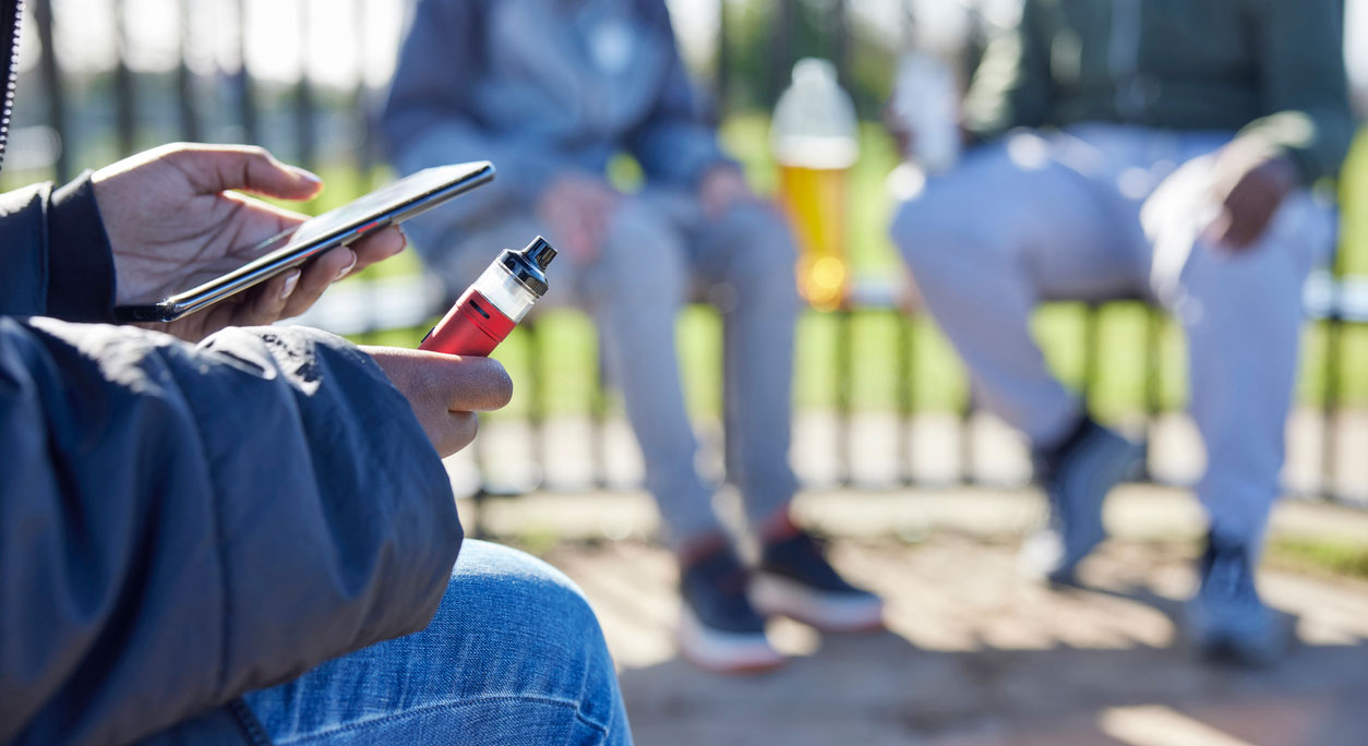 Person sitting on a park bench, holding a vape pen and a phone.