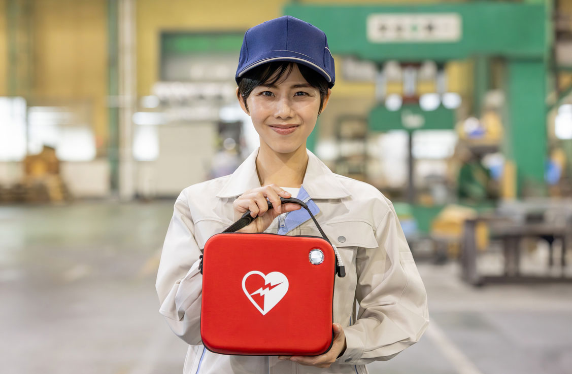 A factory worker smiling and holding the automated external defibrillator kit at her workplace.