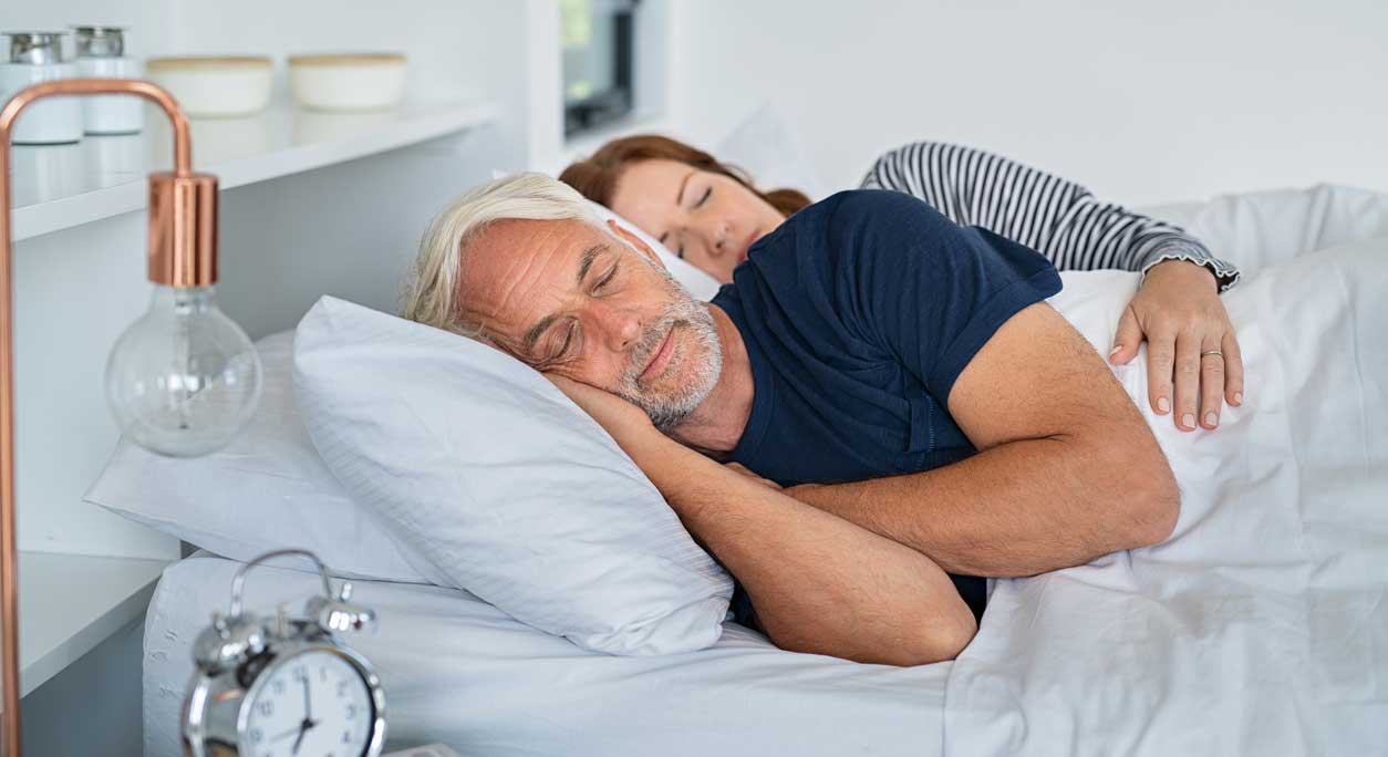A mature couple sleep next to each other in bed with an alarm clock next to them.