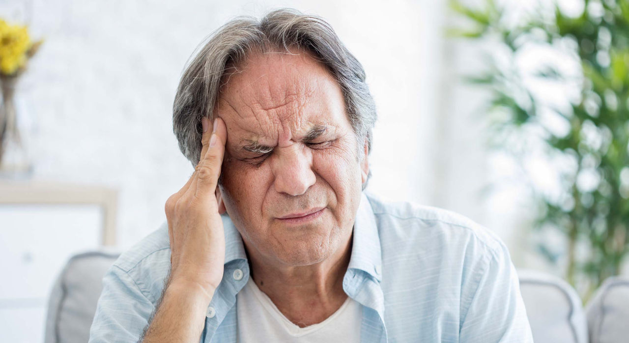 An older man experiencing severe headache pain winces and puts his right hand on his right temple