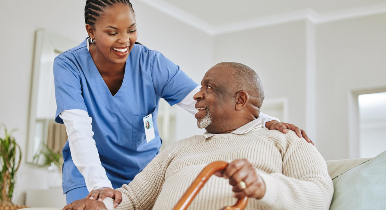 A Black healthcare worker wearing a white long sleeve top under blue scrubs and cornrow braids smiles and checks in on a senior Black man in a beige sweater who is smiling and sitting on a couch while holding a brown cane with his left hand
