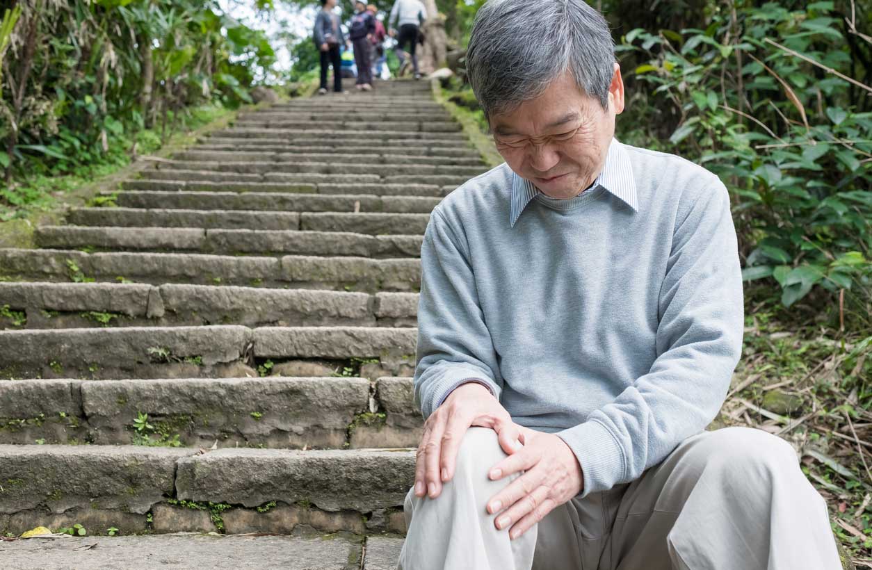 An older man sitting on a stone stairway holds his knee and grimaces in pain.