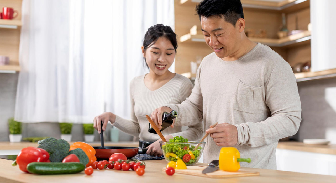 A smiling couple cook together in a bright kitchen, one sauteing a pan and the other drizzling olive oil into a salad bowl. Vegetables and other foods to prevent colorectal cancer are spread on the counter.