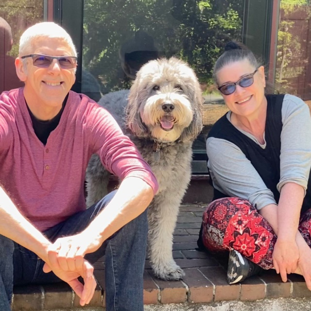 A man sitting on the left, a dog in the middle, and a woman sitting on the right outdoors. The man has white hair with tinted glasses wearing a black undershirt, red long-sleeve top, and jeans. The woman has her hair pulled back and is wearing tinted glasses, a grey long-sleeve top with a black vest on top and a pair of red floral pants.