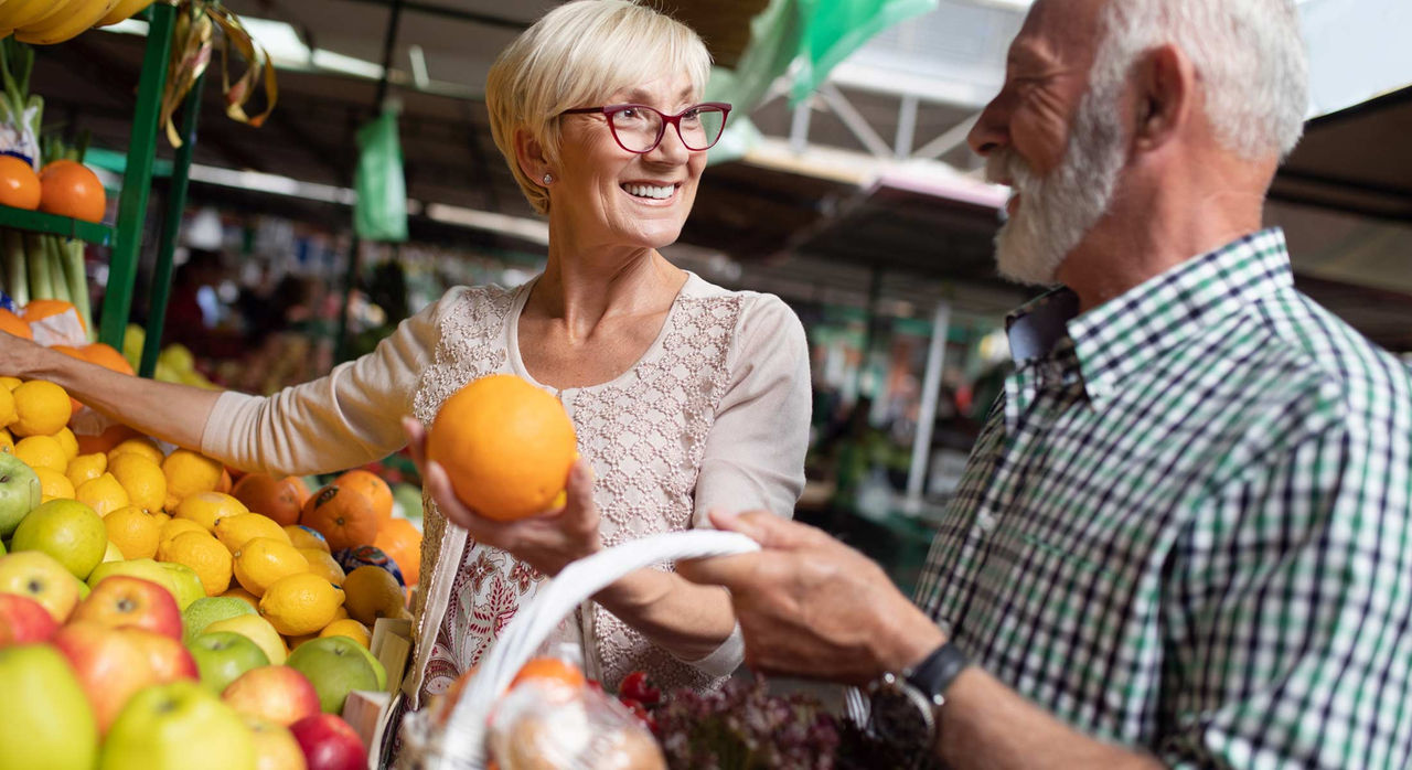 An older white woman with short, light blonde hair smiles at an older white man with short white hair and beard, while they are both shopping for fruits. She reaches her right arm over the fruit stand as the older man smiles back at her as he carries a basket of grapes. The fruit stand at the market has various fruits such as red and green apples, lemons, oranges, and grapefruit.