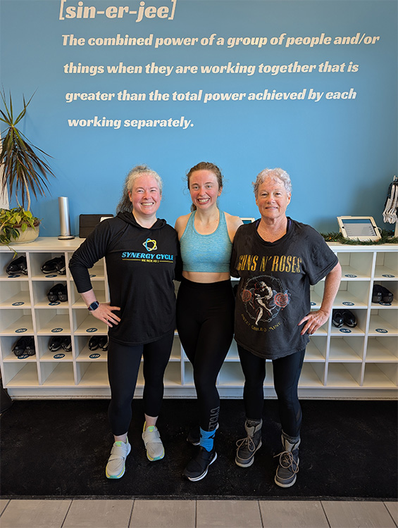 Three women stand together in a gym setting, posing in front of a blue wall with a motivational definition of 'synergy' displayed in large white text.