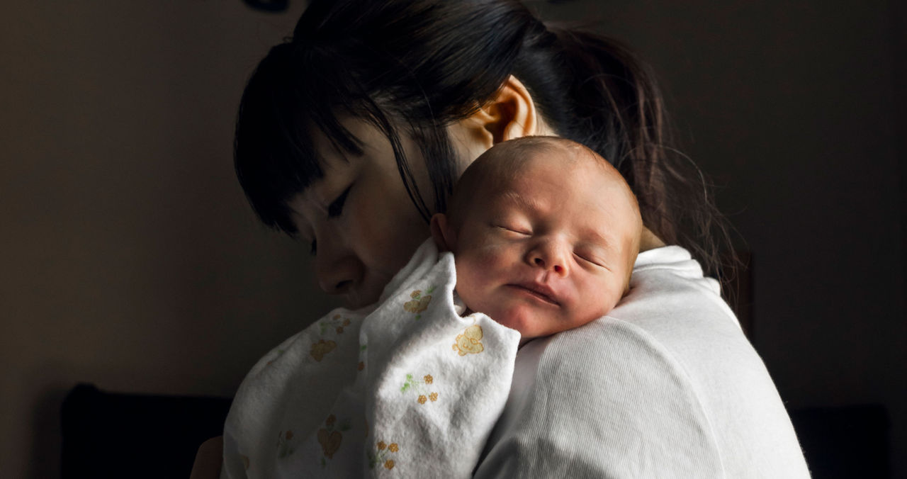 A newborn sleeps on its mother's shoulder in a darkened room.
