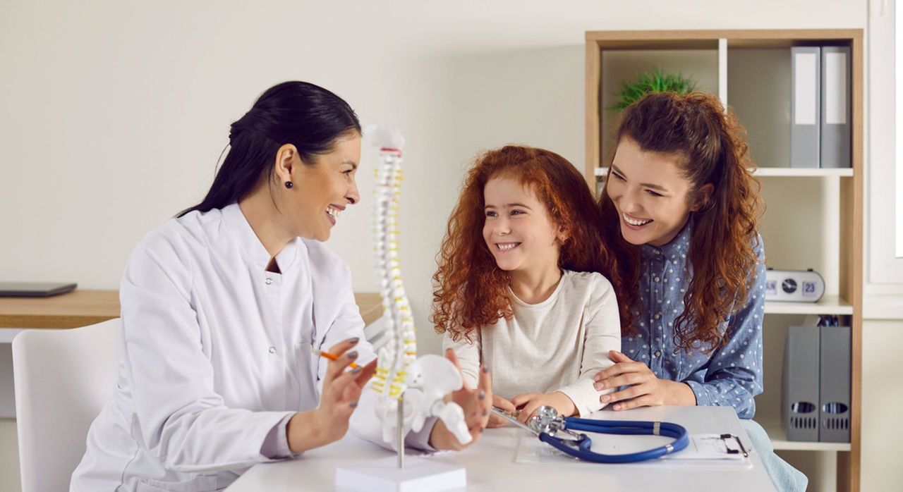 A smiling doctor explains pediatric scoliosis and shows a desk model of a spine to a mother and her daughter.