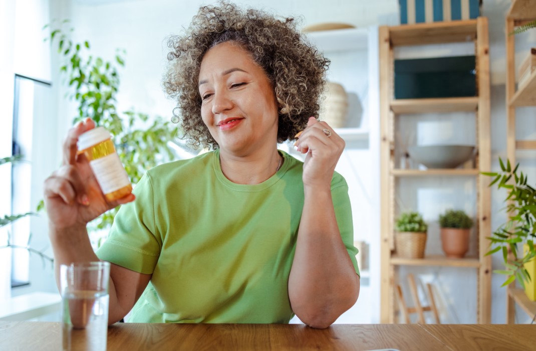 A smiling woman holds a bottle of weight loss medication and reads the label, sitting at a dining table in a bright room with plants and other decor.