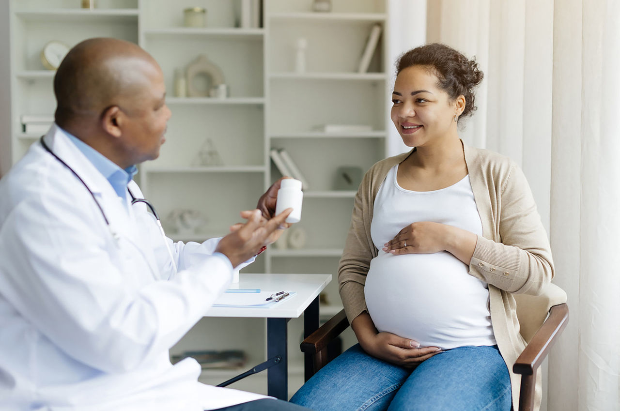 A provider explains safe medications during pregnancy to a pregnant woman in his office, pointing to a pill bottle